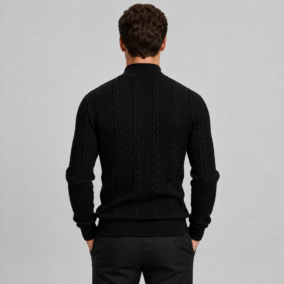 A man viewed from behind wearing a black cable-knit mock-neck sweater and black trousers, standing against a solid light gray studio background.