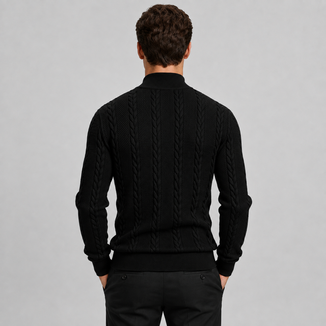 A man viewed from behind wearing a black cable-knit mock-neck sweater and black trousers, standing against a solid light gray studio background.