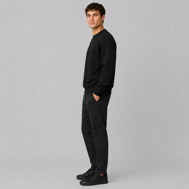 A man wearing a black crewneck sweatshirt, black pants, and black sneakers, standing in profile against a solid light gray background.