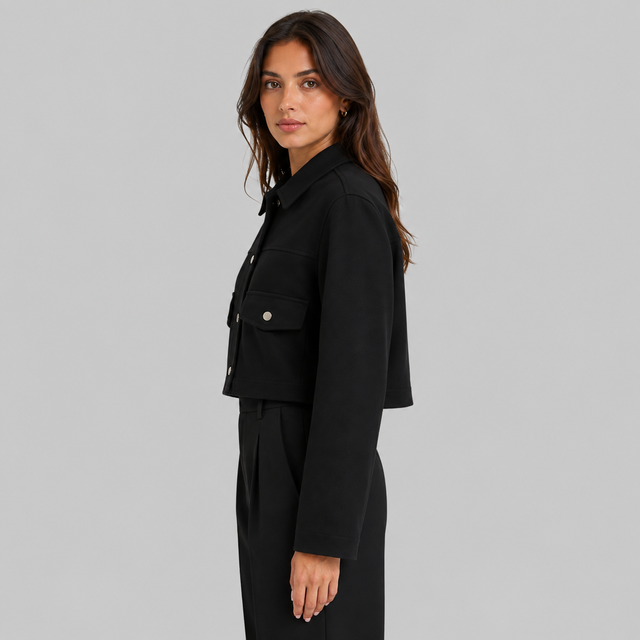 A woman modeling a black cropped jacket with flap chest pockets and silver snap buttons, paired with black trousers, against a solid gray studio background.