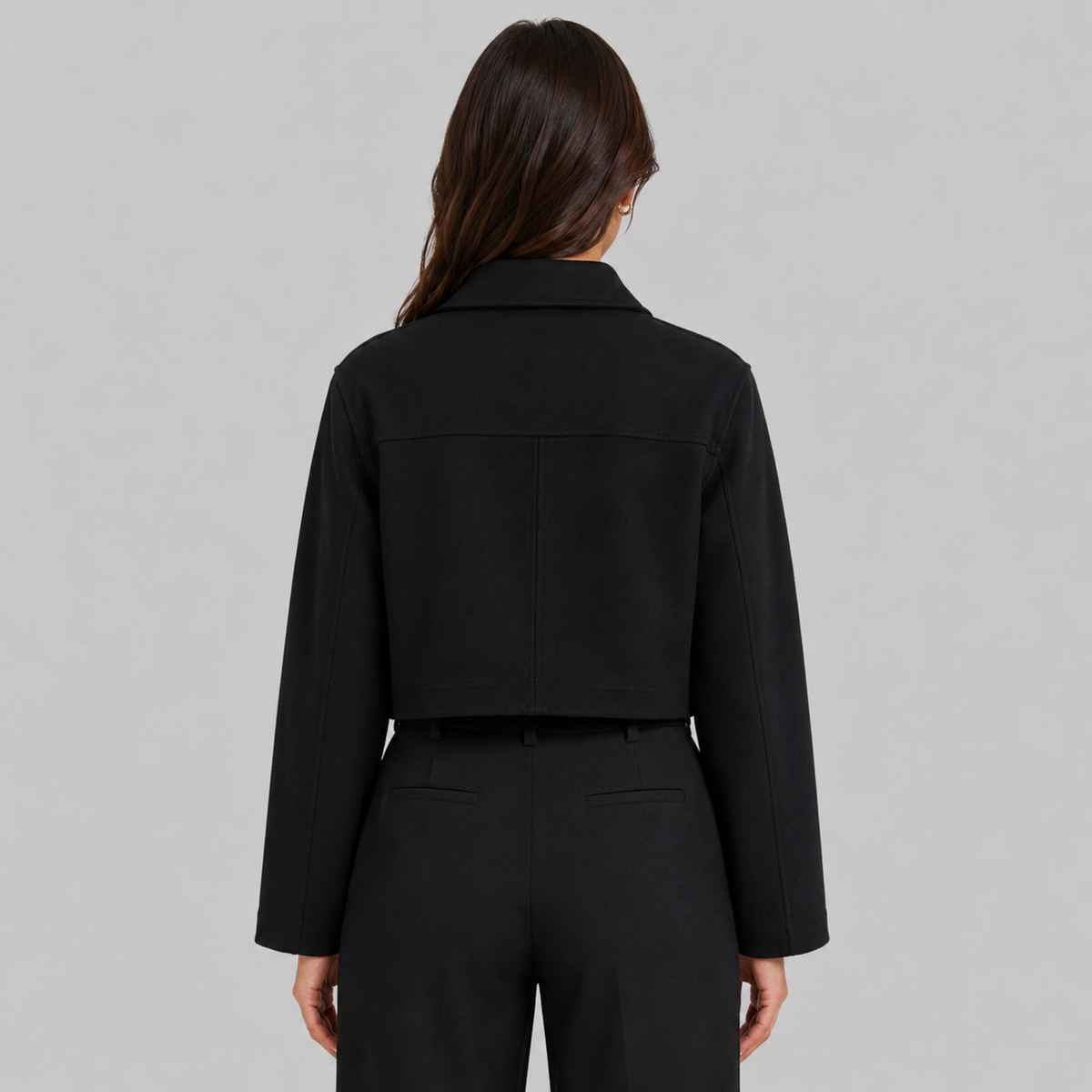 A woman viewed from the back wearing a black cropped jacket with long sleeves and black trousers, against a plain light gray studio background.