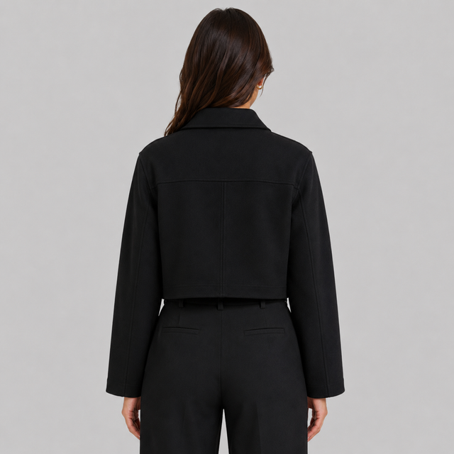 A woman viewed from the back wearing a black cropped jacket with long sleeves and black trousers, against a plain light gray studio background.