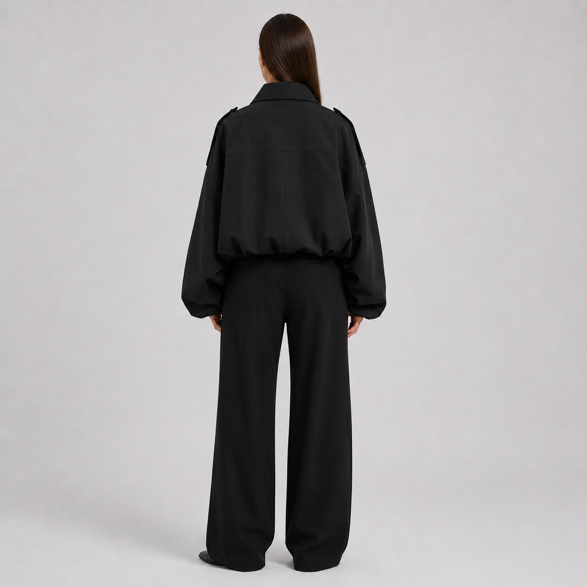 A model viewed from the back wearing a black cropped jacket with wide sleeves and matching black wide-leg trousers, set against a plain light gray studio background.