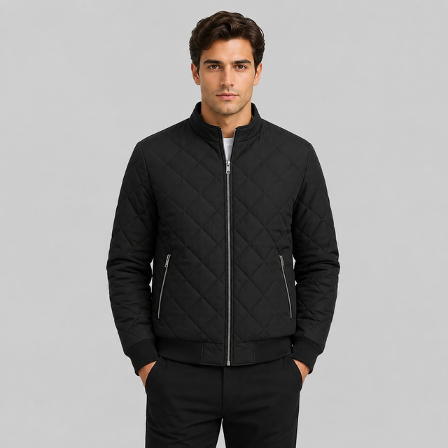 A man wearing a black diamond-quilted zip-up bomber jacket with silver zippers and a stand-up collar, paired with black trousers, against a solid gray studio background.