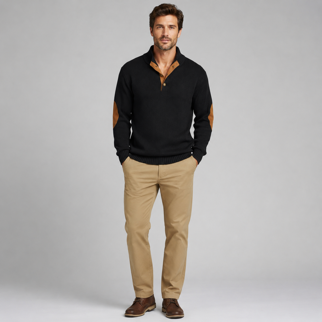 A man modeling a black knit polo sweater with tan elbow patches and a tan inner placket, paired with khaki pants and brown shoes, against a light gray studio background.