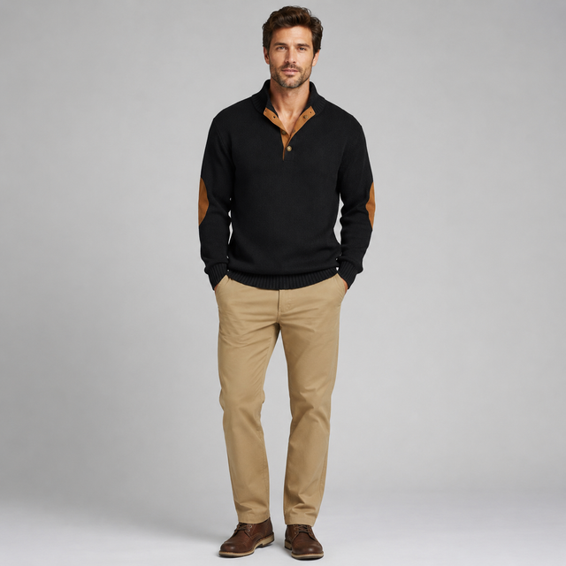 A man modeling a black knit polo sweater with tan elbow patches and a tan inner placket, paired with khaki pants and brown shoes, against a light gray studio background.