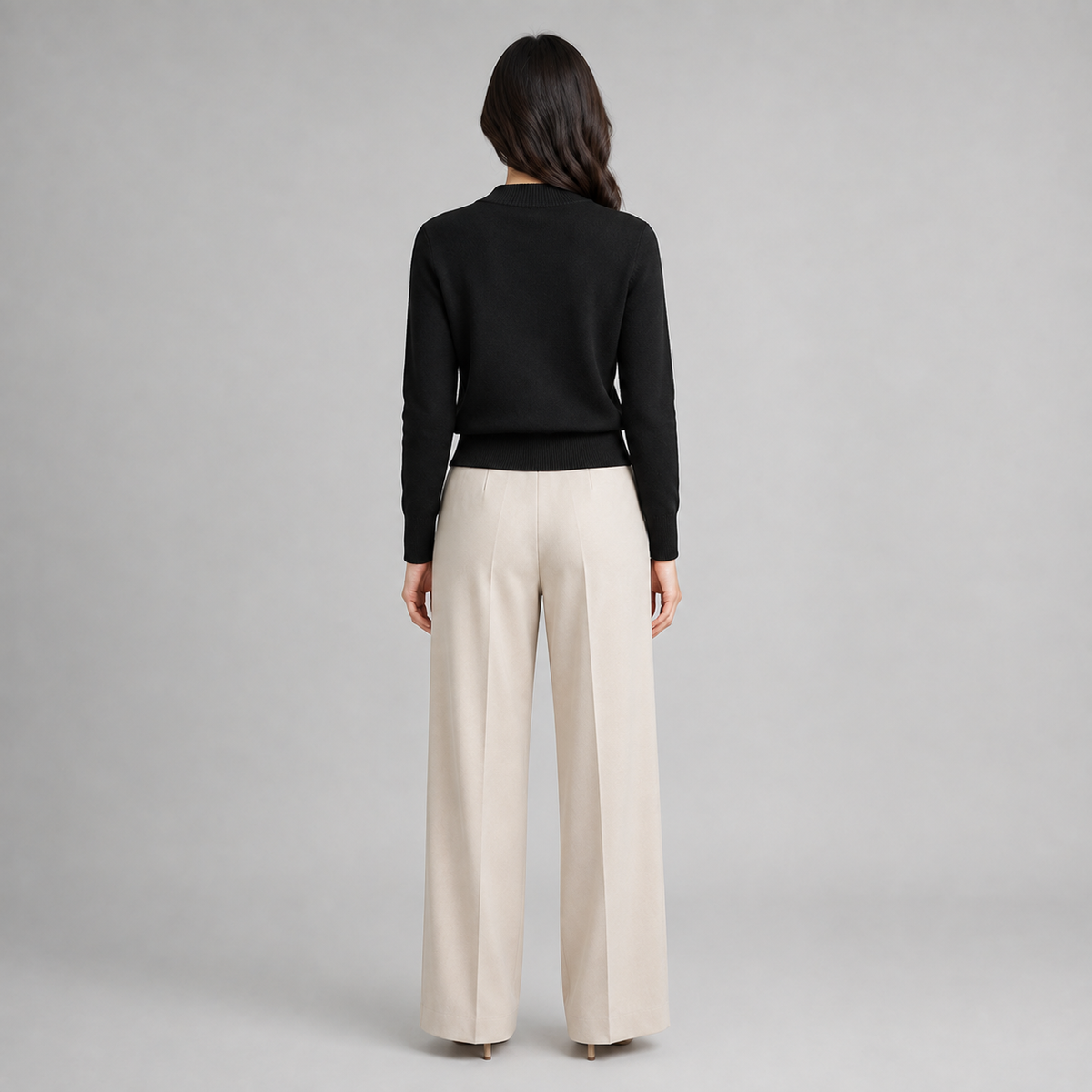 A woman shown from the back wearing a black long-sleeve knit top and light beige wide-leg trousers, standing against a neutral gray studio background.