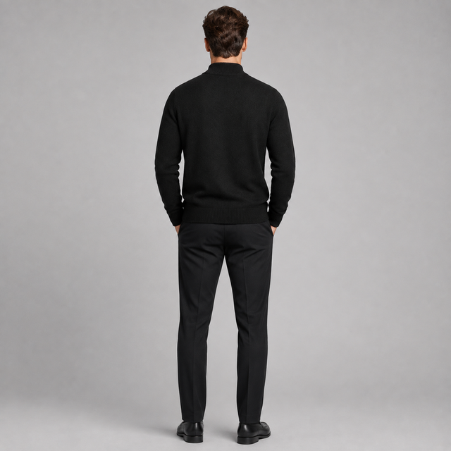 A man viewed from the back wearing a black mock-neck knit sweater and black trousers, standing against a neutral gray studio background.