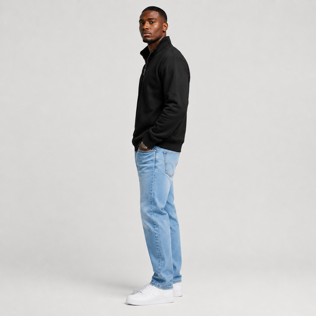 A man standing in profile wearing a black quarter-zip pullover, light blue jeans, and white sneakers against a light gray studio background.