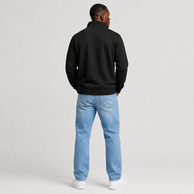 A man viewed from behind wearing a black quarter-zip sweatshirt, light blue jeans, and white sneakers, standing against a light gray studio background.