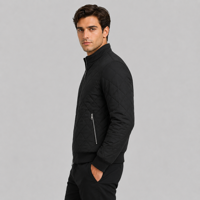 A man wearing a black quilted zip-up jacket with a stand collar and a diagonal zip pocket on the chest, paired with black trousers, photographed against a solid light gray background.