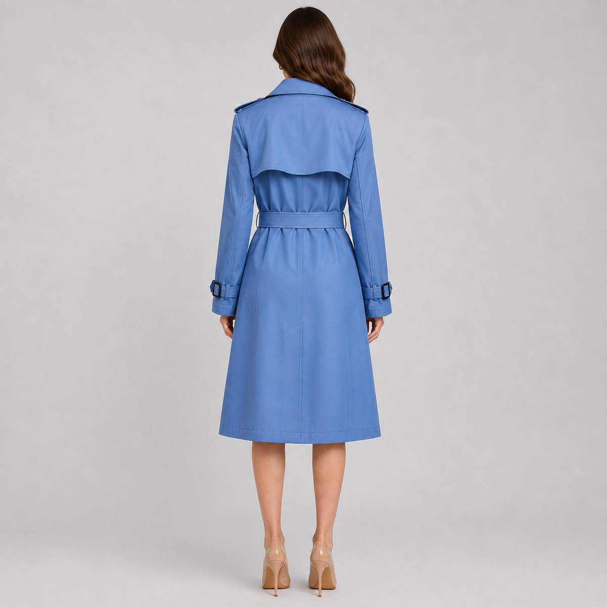 A woman viewed from the back wearing a blue trench coat with a matching waist belt, paired with beige high heels, against a light gray studio background.