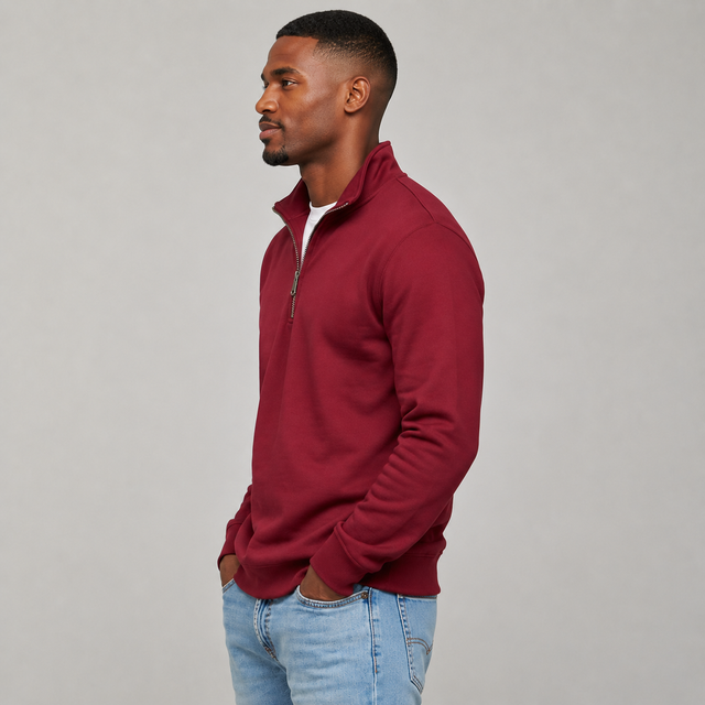 A man in profile wearing a burgundy quarter-zip pullover sweater over a white undershirt and light blue jeans, standing against a neutral gray studio background.
