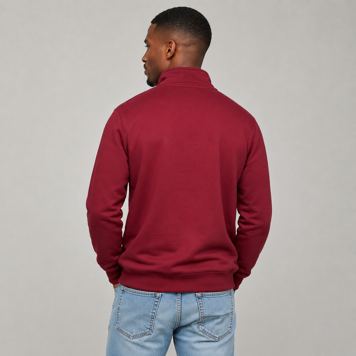 A man viewed from the back wearing a burgundy quarter-zip sweatshirt and light blue jeans, standing against a neutral gray studio background.