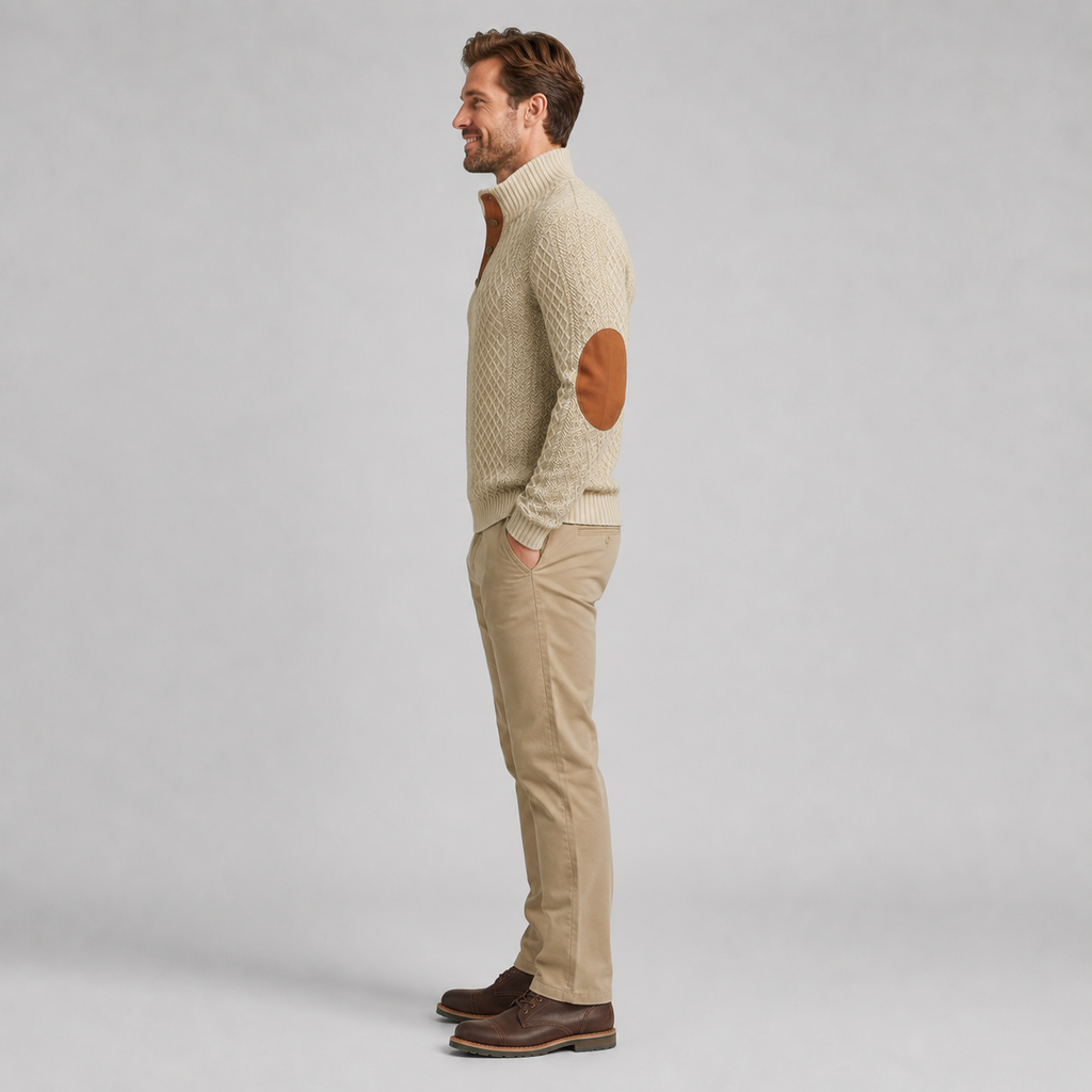 A man in side profile wearing a cream cable-knit sweater with brown elbow patches, beige trousers, and dark brown shoes, standing against a light gray studio background.