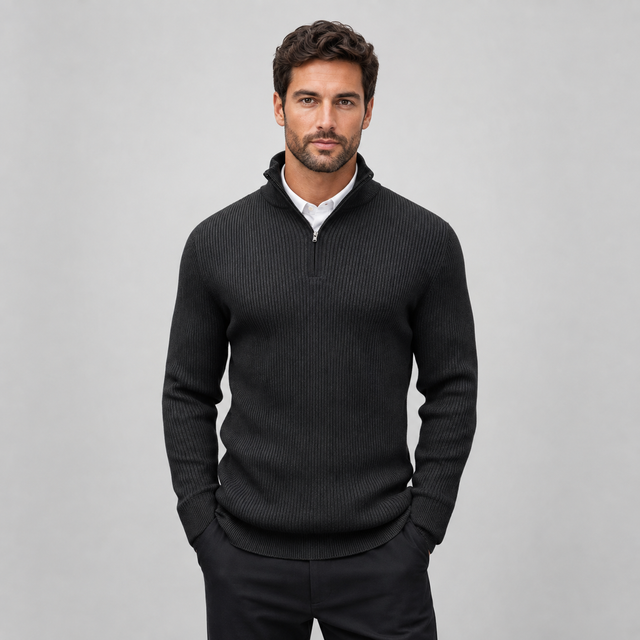 A man wearing a dark charcoal ribbed quarter-zip sweater over a white collared shirt, standing against a light gray studio background.
