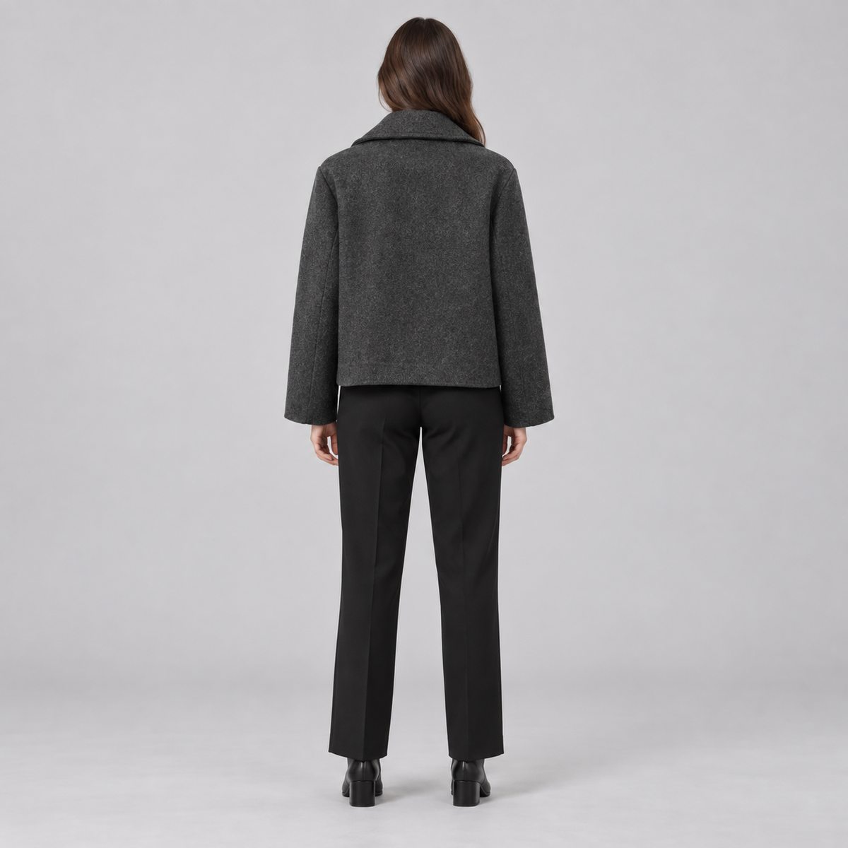 A woman viewed from the back wearing a dark gray cropped jacket and black trousers against a light gray studio background.