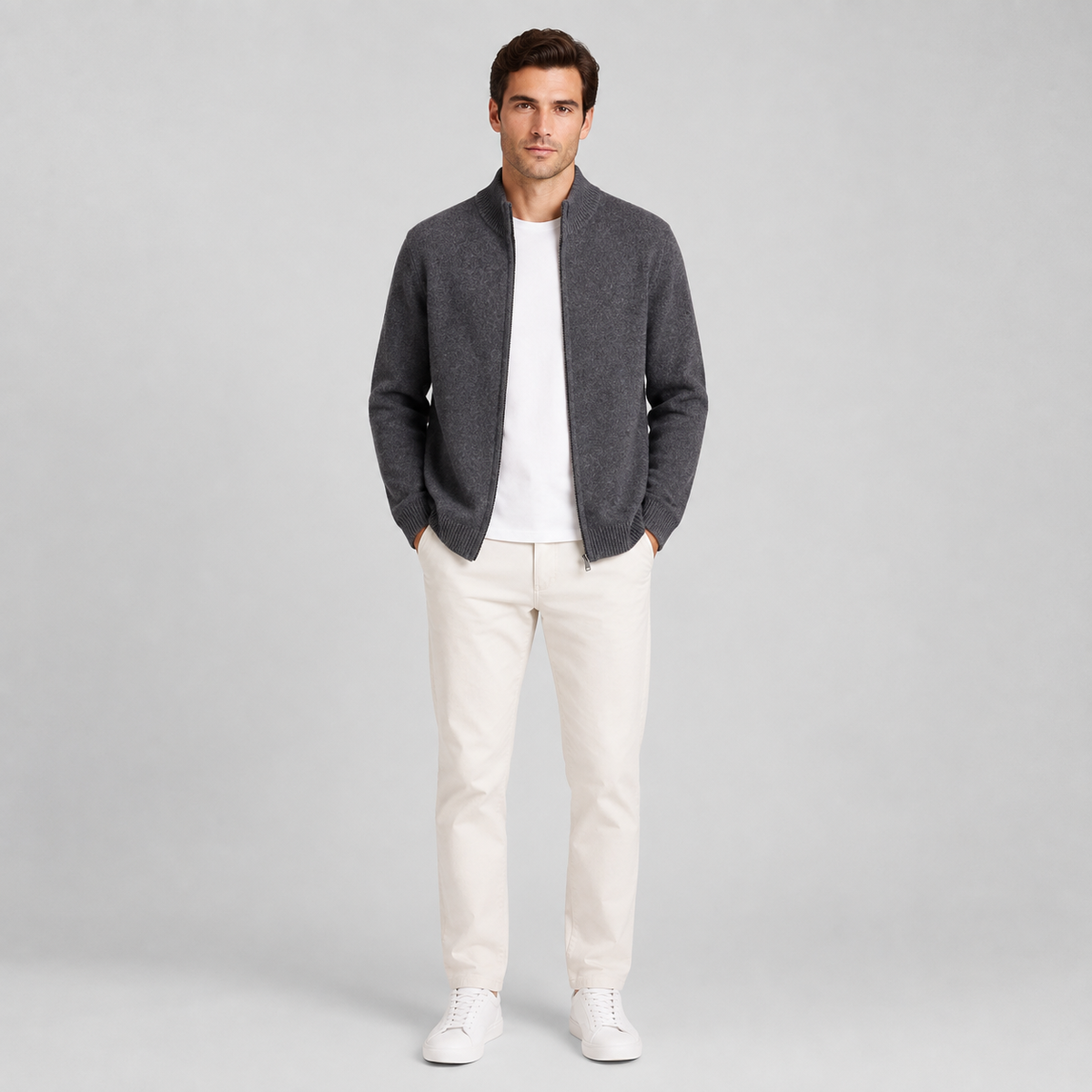 A man modeling a dark gray knit zip-up cardigan over a white t-shirt, paired with off-white trousers and white sneakers, against a light gray studio background.