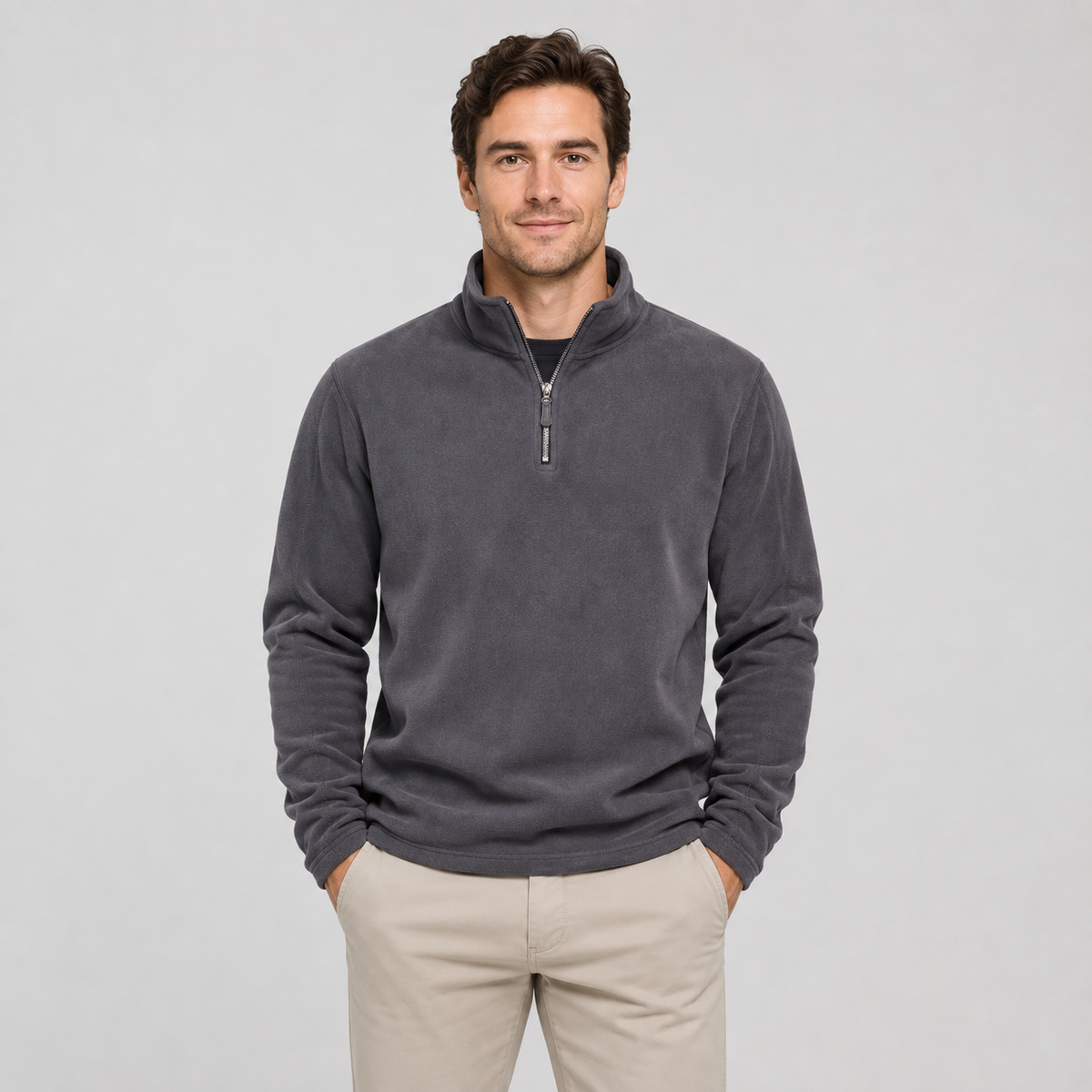 A man wearing a dark gray quarter-zip fleece pullover sweater and beige pants, standing against a light gray studio background.
