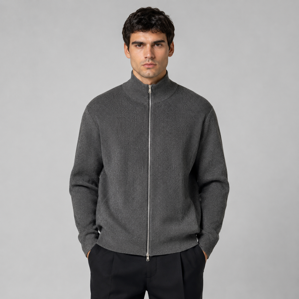 A man wearing a dark gray ribbed knit zip-up mock neck sweater, paired with black trousers, standing against a solid gray studio background.