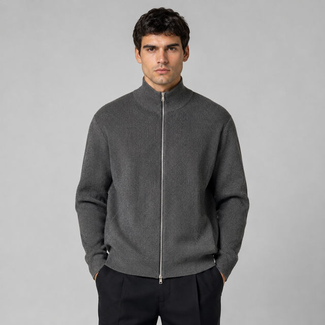 A man wearing a dark gray ribbed knit zip-up mock neck sweater, paired with black trousers, standing against a solid gray studio background.