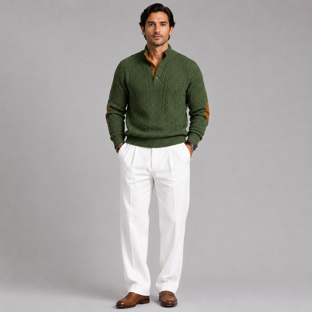 A man modeling a dark green cable-knit sweater with brown elbow patches, paired with white trousers and brown leather shoes, against a solid gray studio background.