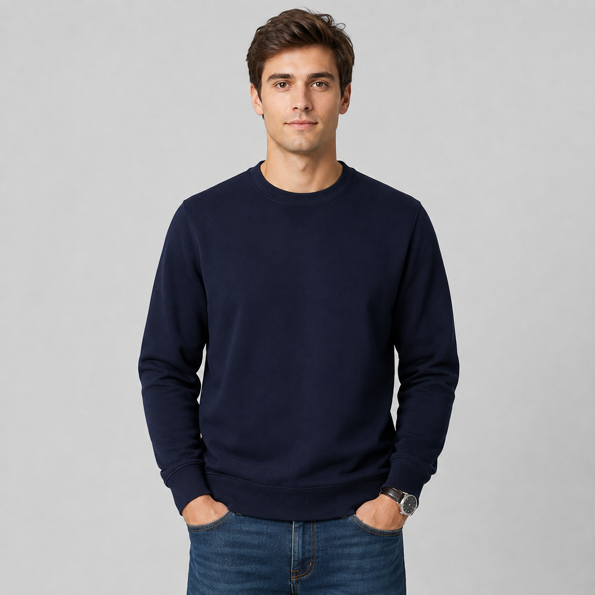 A man wearing a dark navy blue crewneck sweatshirt and blue jeans, standing against a light gray studio background.