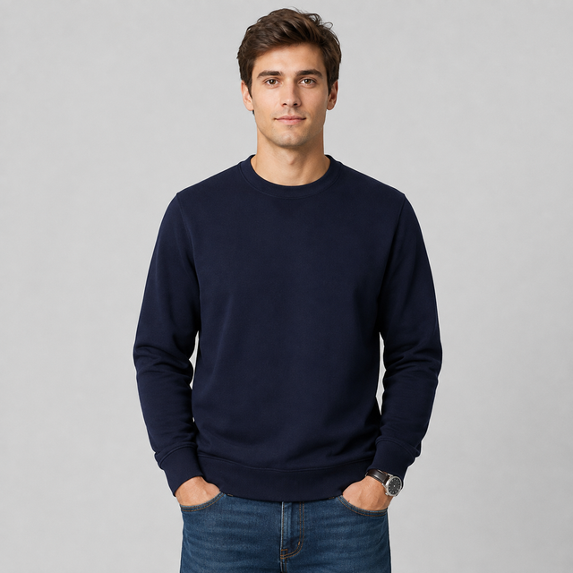 A man wearing a dark navy blue crewneck sweatshirt and blue jeans, standing against a light gray studio background.