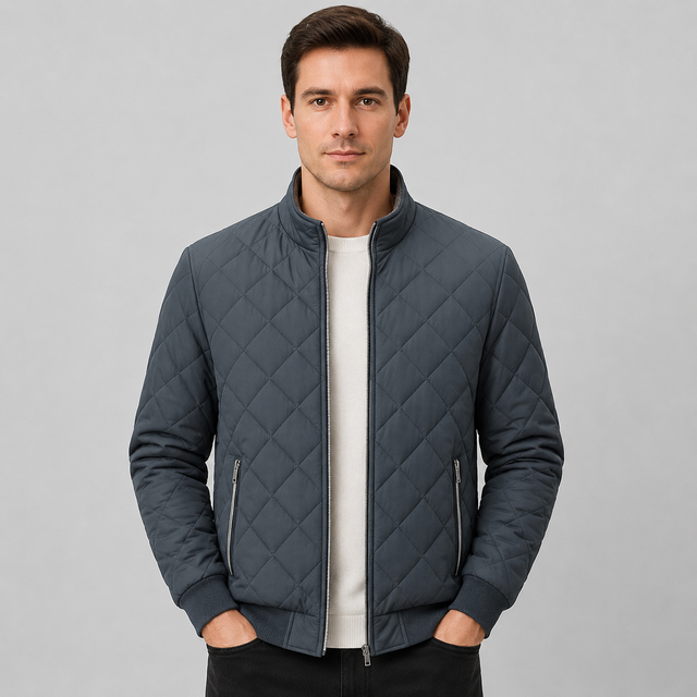 A man wearing a dark slate gray diamond-quilted bomber jacket over a white crewneck shirt, standing against a neutral light gray background.