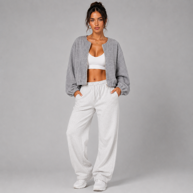 A woman modeling a gray cropped knit cardigan over a white sports bra and white sweatpants, standing against a plain light gray studio background.