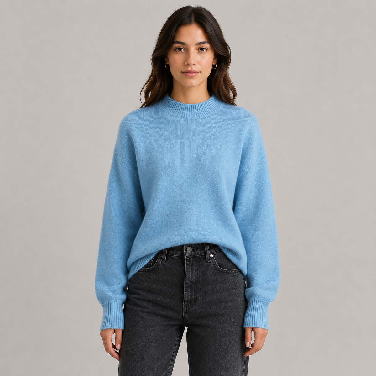 A woman wearing a light blue crewneck knit sweater and dark gray jeans, standing against a neutral light gray studio background.