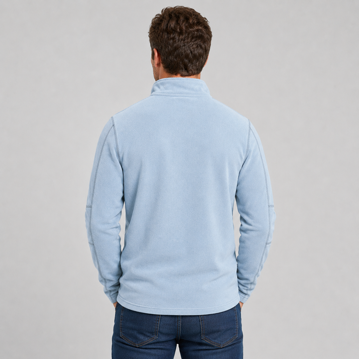 A man viewed from behind wearing a light blue long-sleeve quarter-zip fleece jacket and dark blue jeans, standing against a neutral light gray background.