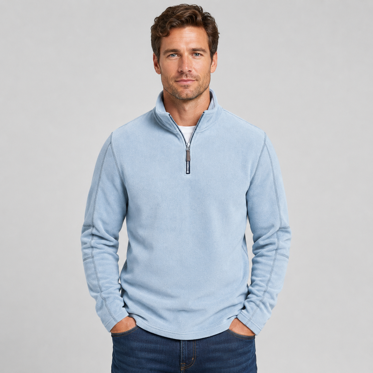 A man wearing a light blue quarter-zip fleece pullover sweater and dark blue jeans, standing against a neutral light gray background.