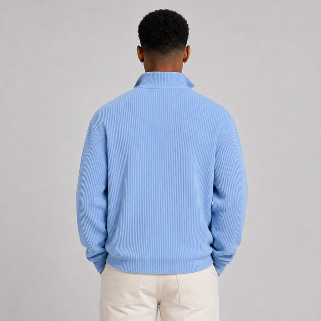A man viewed from behind wearing a light blue ribbed knit quarter-zip sweater and cream pants, standing against a neutral gray studio background.