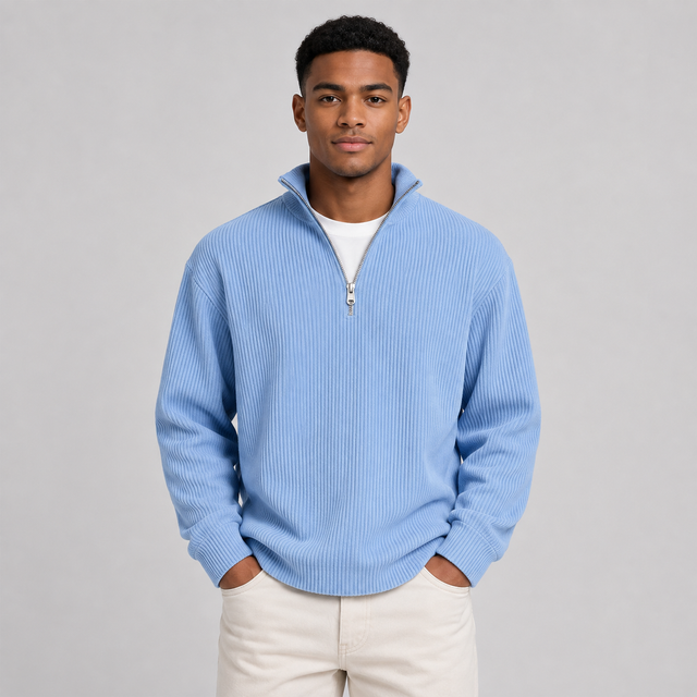 A man wearing a light blue ribbed quarter-zip sweater over a white t-shirt and cream pants, standing against a neutral gray studio background.