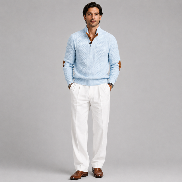 A man modeling a light blue textured quarter-zip sweater with brown leather elbow patches, paired with white trousers and brown leather shoes, against a solid gray studio background.