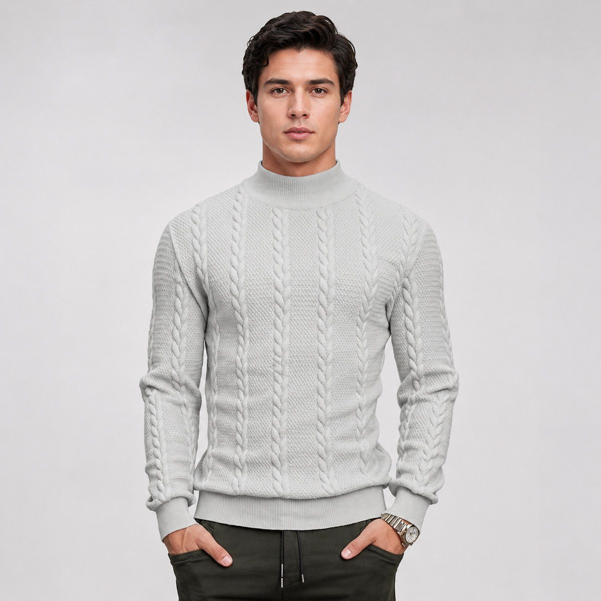 A man wearing a light gray cable-knit mock neck sweater, paired with dark pants, standing against a light gray studio background.