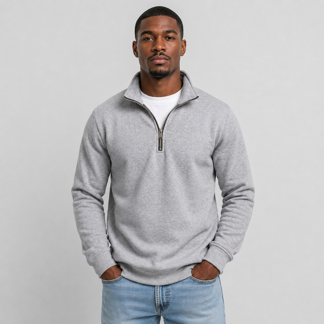 A man wearing a light gray heathered quarter-zip sweatshirt over a white t-shirt and blue jeans, standing against a light gray studio background.