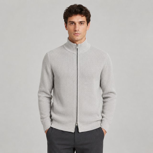 A man wearing a light gray ribbed knit zip-up mock neck sweater and dark gray trousers, standing against a solid light gray studio background.