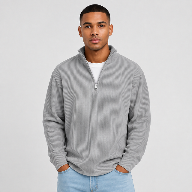 A man wearing a light gray ribbed quarter-zip pullover sweater over a white t-shirt and light blue jeans, standing against a light gray studio background.