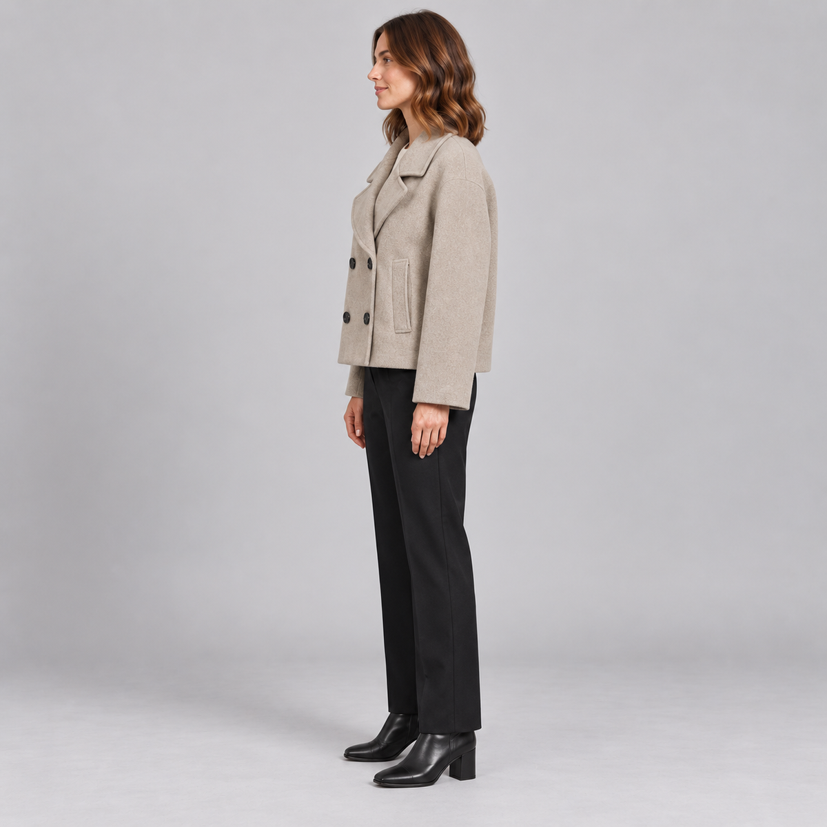 A woman stands in profile wearing a light beige double-breasted blazer, black trousers, and black ankle boots against a plain light gray studio background.