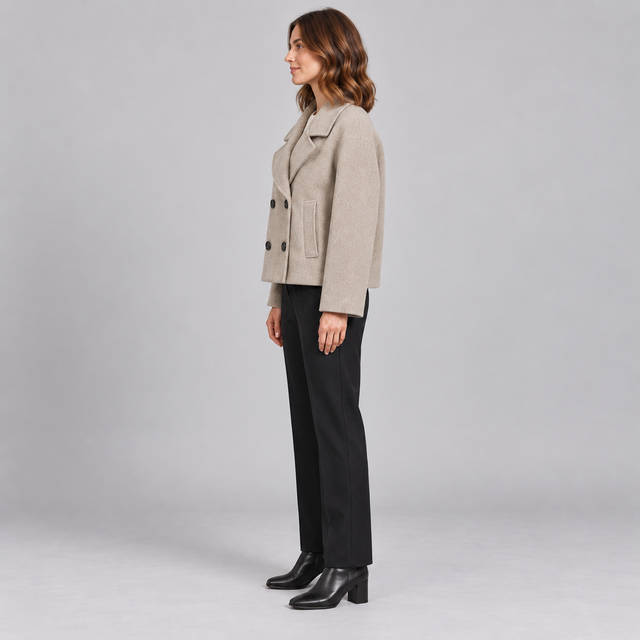 A woman stands in profile wearing a light beige double-breasted blazer, black trousers, and black ankle boots against a plain light gray studio background.