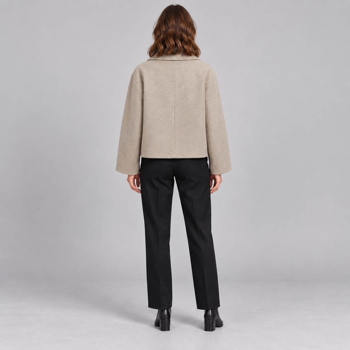 A woman shown from the back wearing a cropped beige jacket with long sleeves, black trousers, and black ankle boots, standing against a light gray studio background.