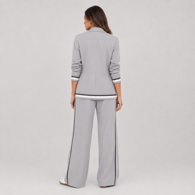 A woman shown from the back wearing a light gray two-piece tracksuit with black and white striped accents on the cuffs, hem, and side seams, against a neutral light gray studio background.