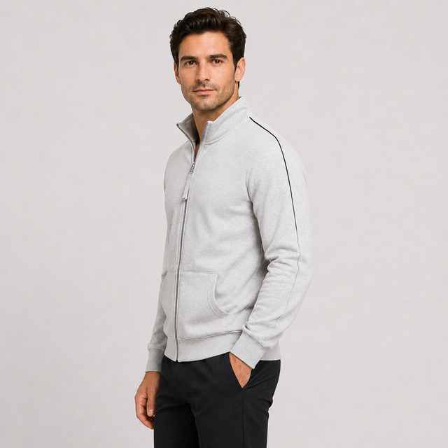 A man wearing a light gray zip-up track jacket with black piping details on the shoulders and sleeves, paired with black pants, photographed against a neutral light gray background.