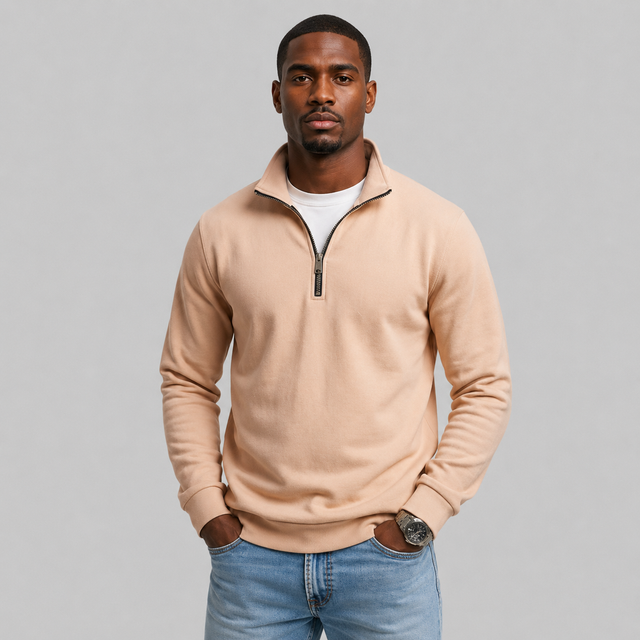 A man wearing a light peach-beige quarter-zip sweatshirt over a white t-shirt and blue jeans, standing against a solid gray studio background.