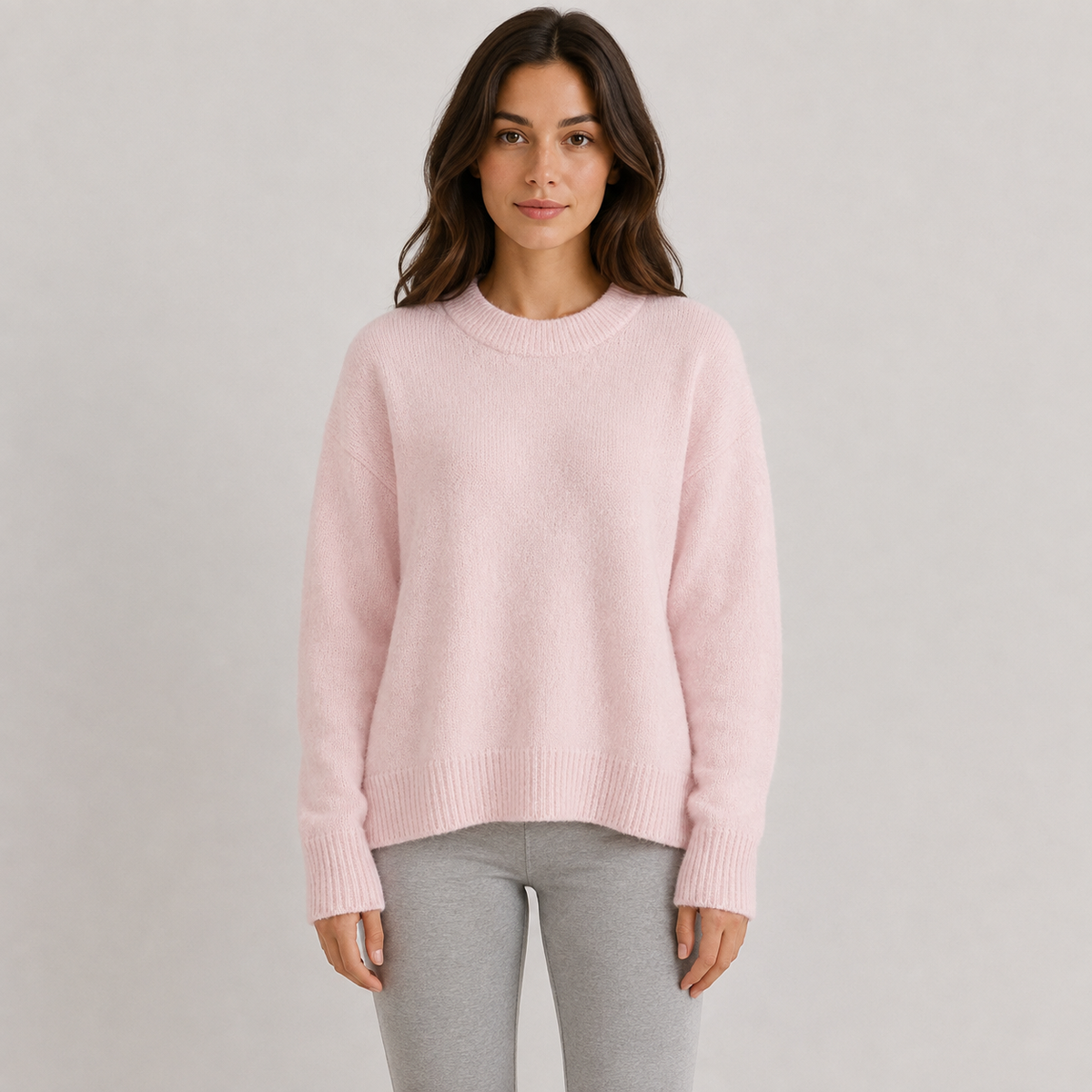 A woman wearing a light pink crewneck knit sweater and light gray leggings, standing against a neutral light gray studio background.