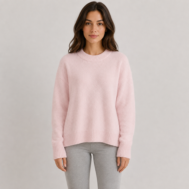 A woman wearing a light pink crewneck knit sweater and light gray leggings, standing against a neutral light gray studio background.