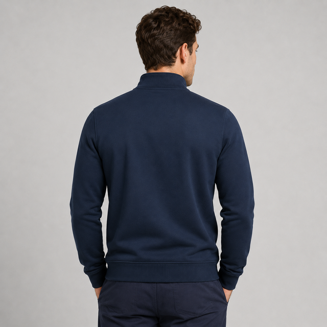 A man viewed from behind wearing a navy blue mock-neck or high-collar long-sleeve top and matching navy pants against a plain light gray studio background.