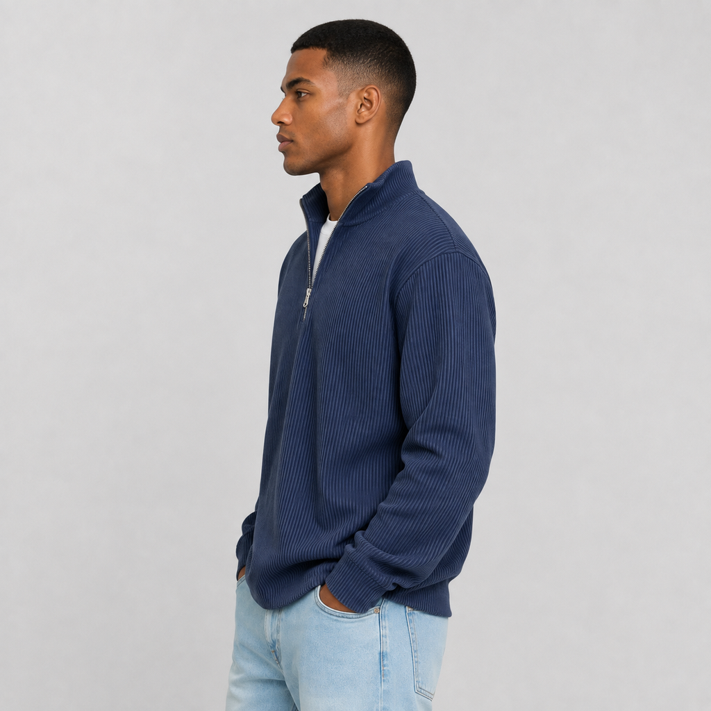 A man in profile wearing a navy blue ribbed quarter-zip pullover sweater over a white t-shirt and light blue jeans, standing against a neutral light gray background.
